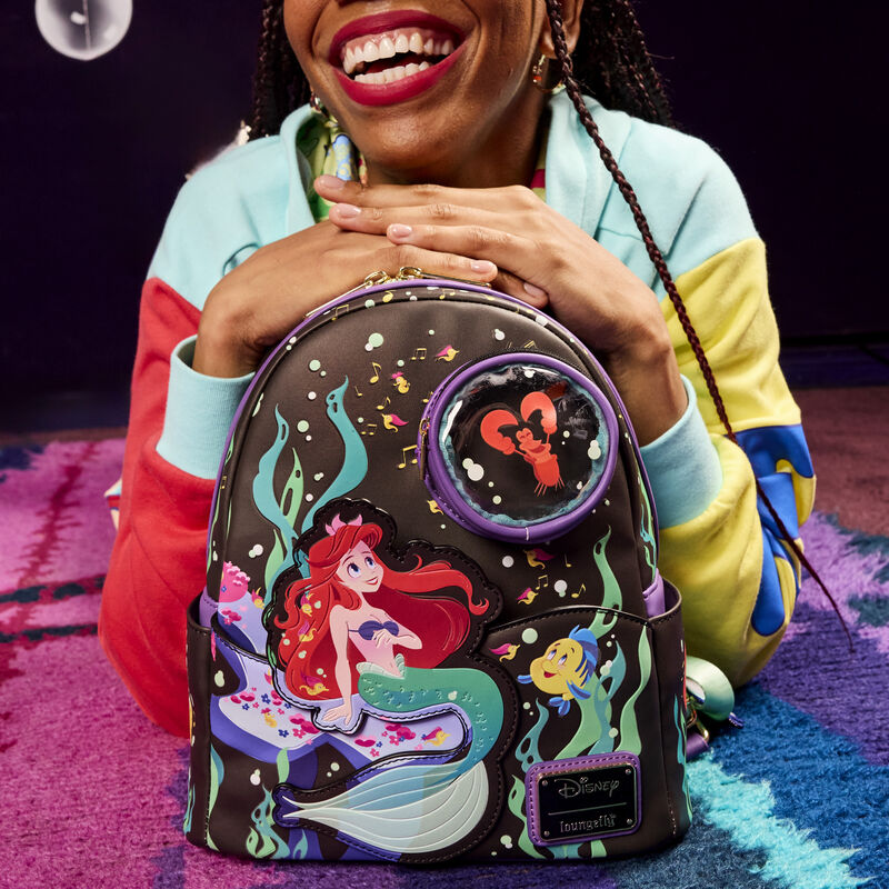 Smiling woman laying on her stomach on a carpet with her hands resting on the top of the Little Mermaid Life in the Bubbles Mini Backpack